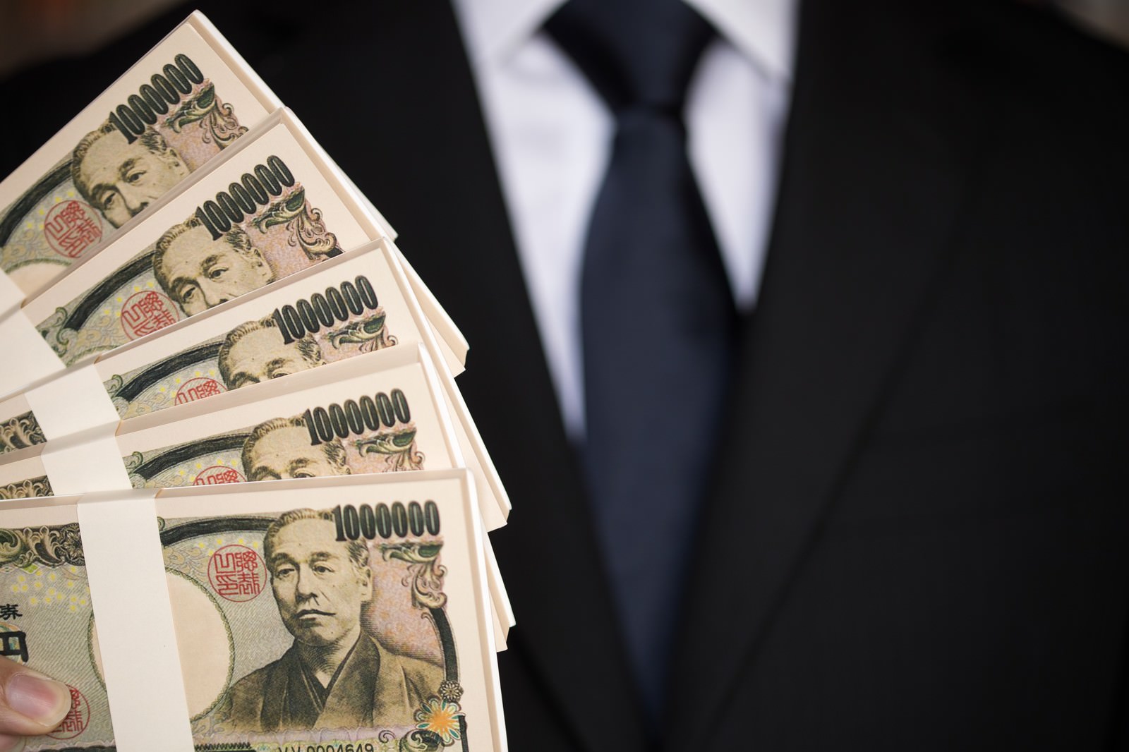 黒いスーツを着た男性が1万円札の札束を扇状に広げて手に持っている様子