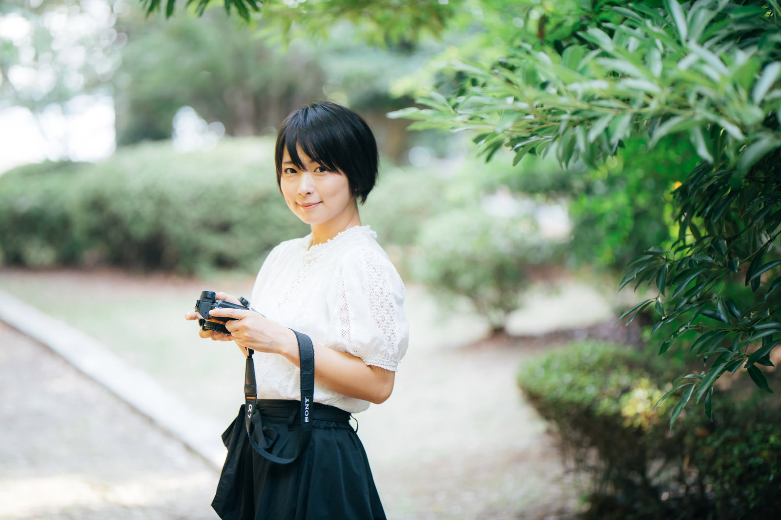 白いトップスを着た女性が黒いカメラを持ちながら笑顔で会釈している様子［モデル：にゃるる］