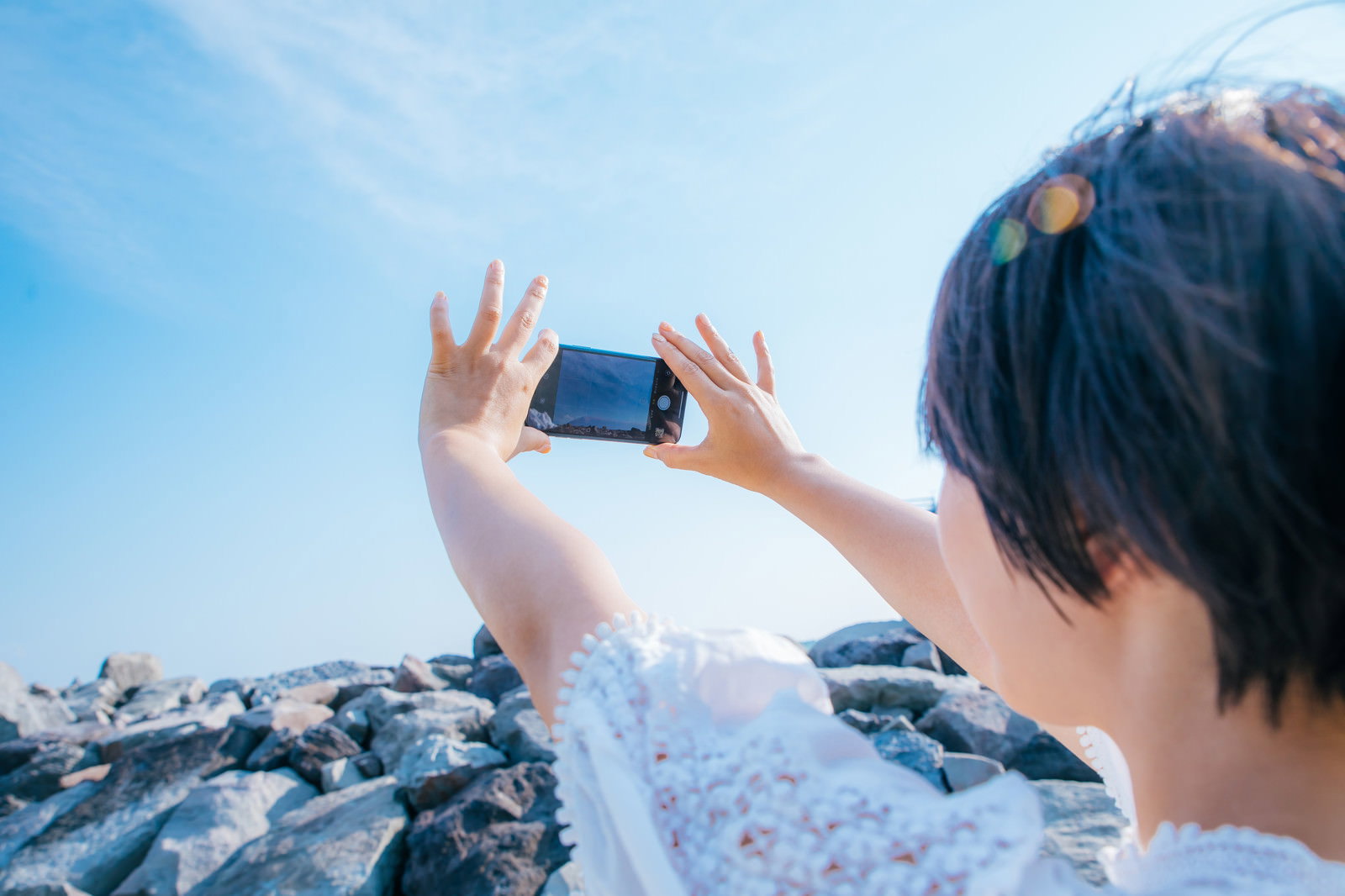青空に向かってスマートフォンを掲げて撮影している女性の後ろ姿［モデル：にゃるる］