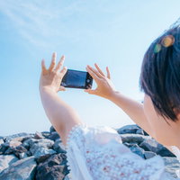 スマホで青空を撮影する女性の後ろ姿の写真
