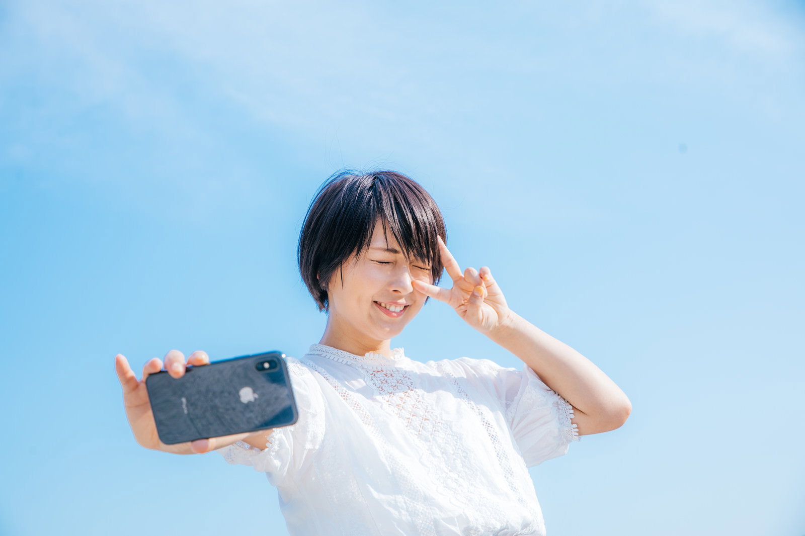 青空を背景にスマートフォンを持ちピースサインをする笑顔の女性［モデル：にゃるる］