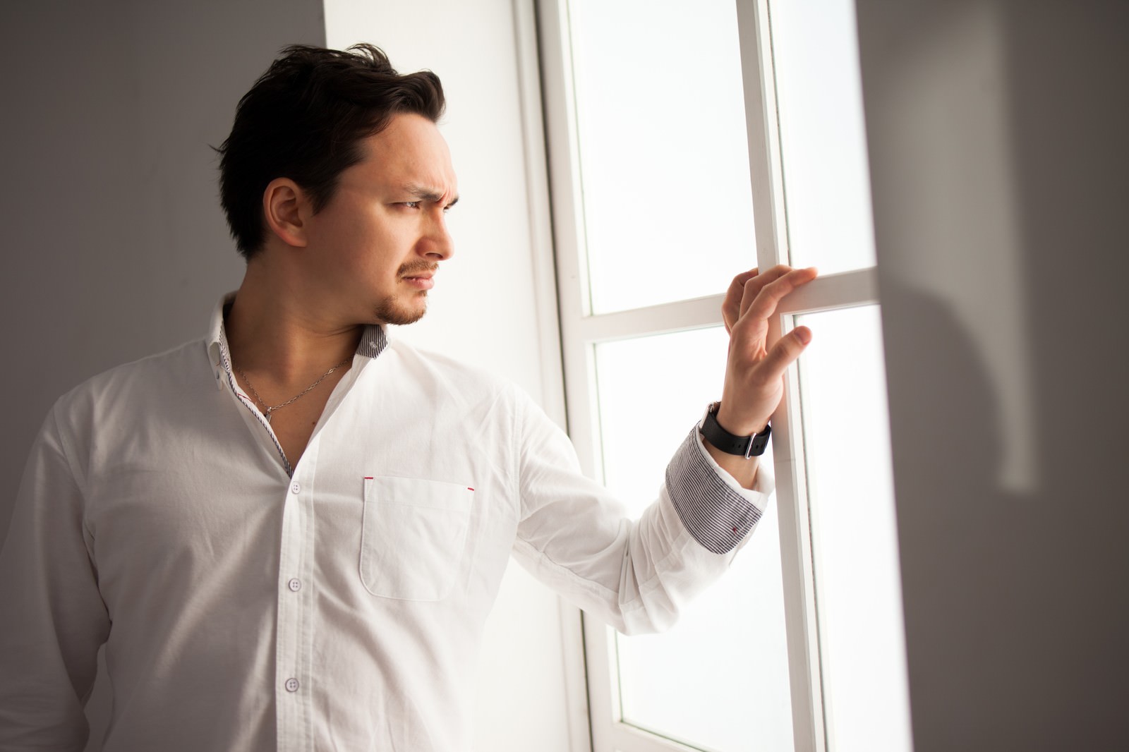 白いシャツの男性が窓に手をかけ、警戒した表情で窓外を見つめている［モデル：Max_Ezaki］