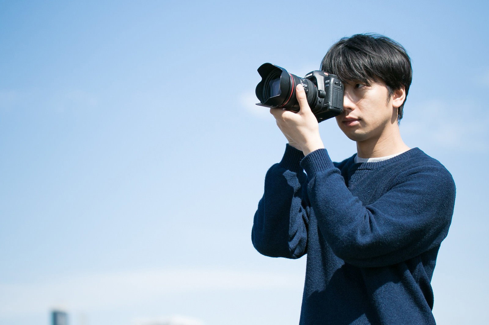 紺色のセーターを着た男性が青空の下で一眼レフカメラを構えて撮影している［モデル：大川竜弥］