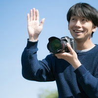 カメラを手に笑顔で手を振るカメラ男子、紺色セーター姿の写真