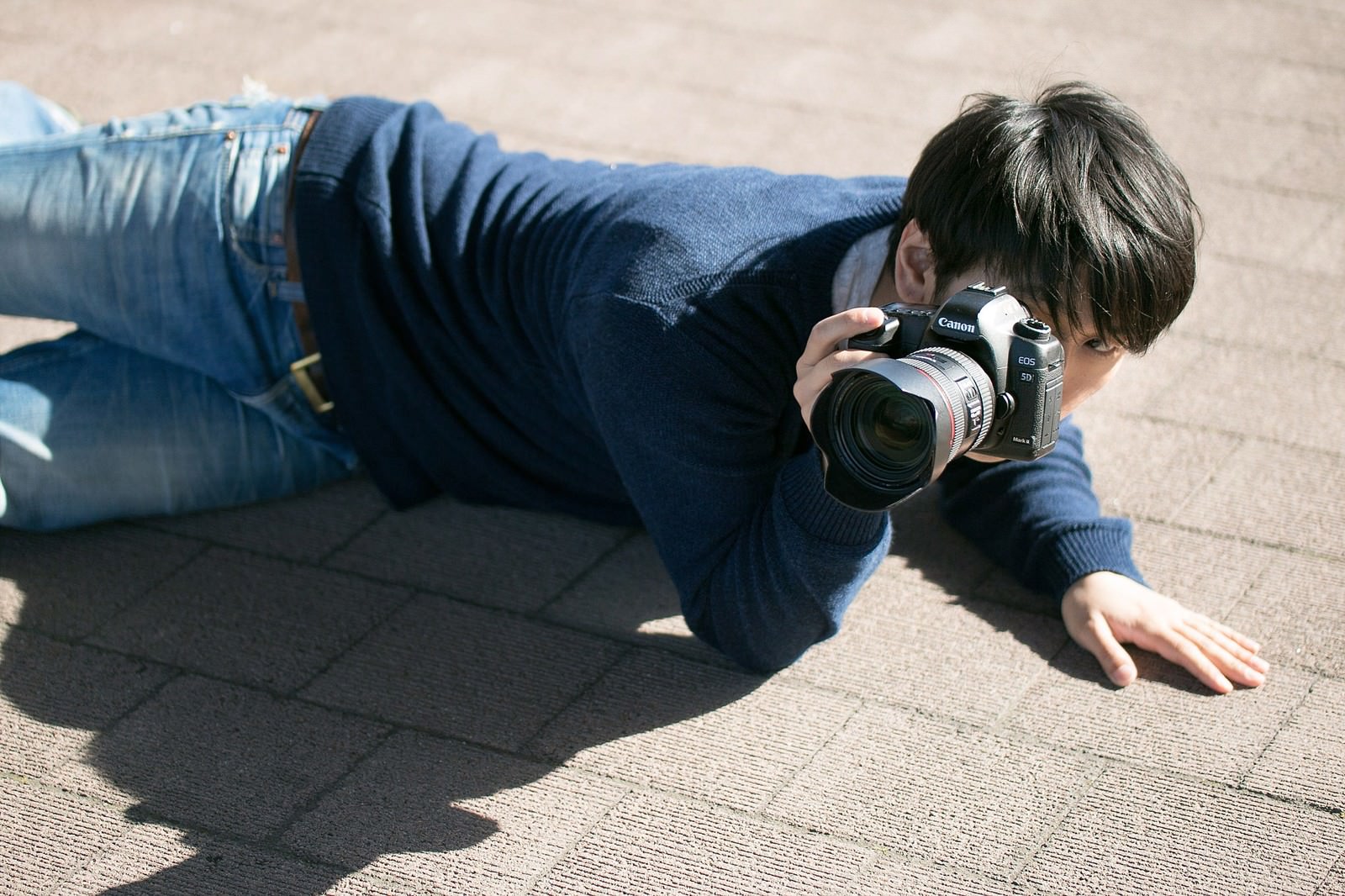 地面に腹ばいでCanon一眼レフを構える男性カメラマン［モデル：大川竜弥］