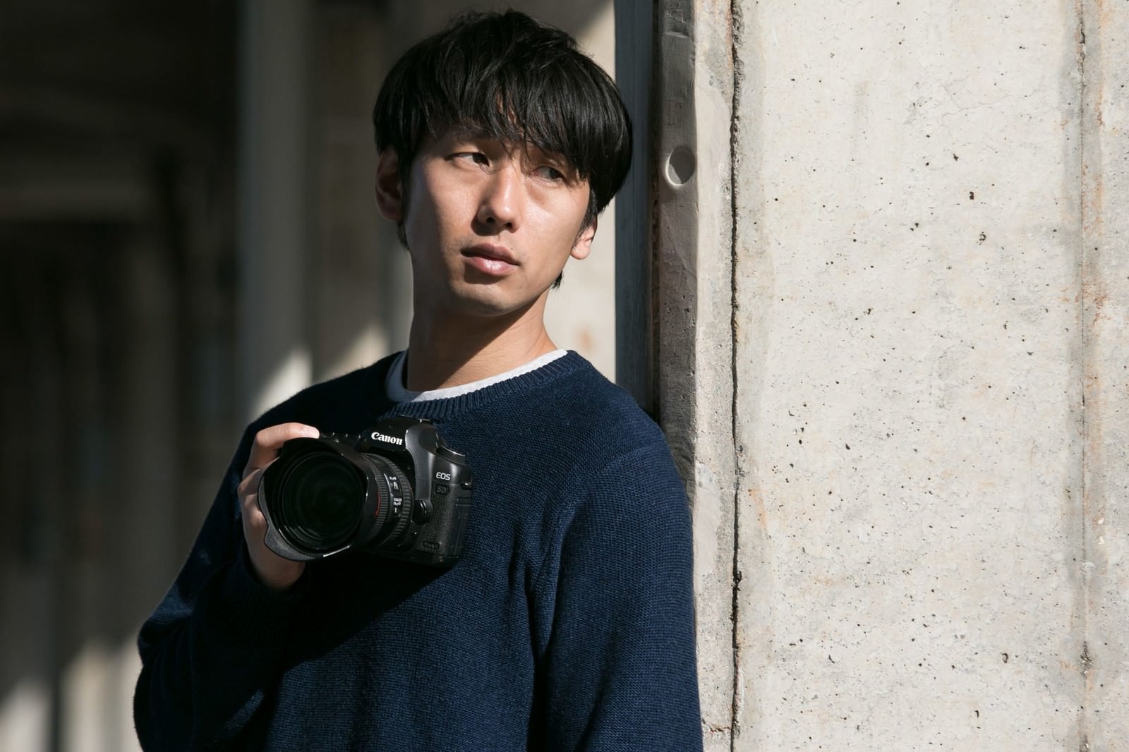 柱に寄り添い一眼レフカメラを構える男性カメラマン［モデル：大川竜弥］