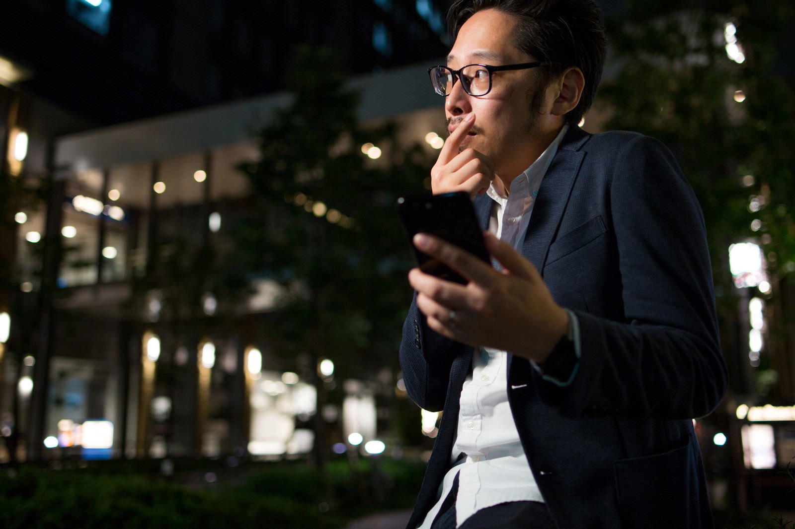 夜の街でスマホのマップを見て考える眼鏡の男性［モデル：ゆうせい］