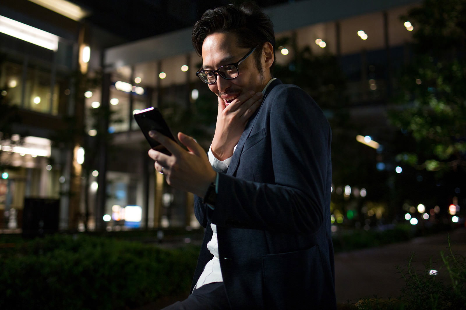 夜の屋外でスマホを見てニヤける眼鏡の男性［モデル：ゆうせい］