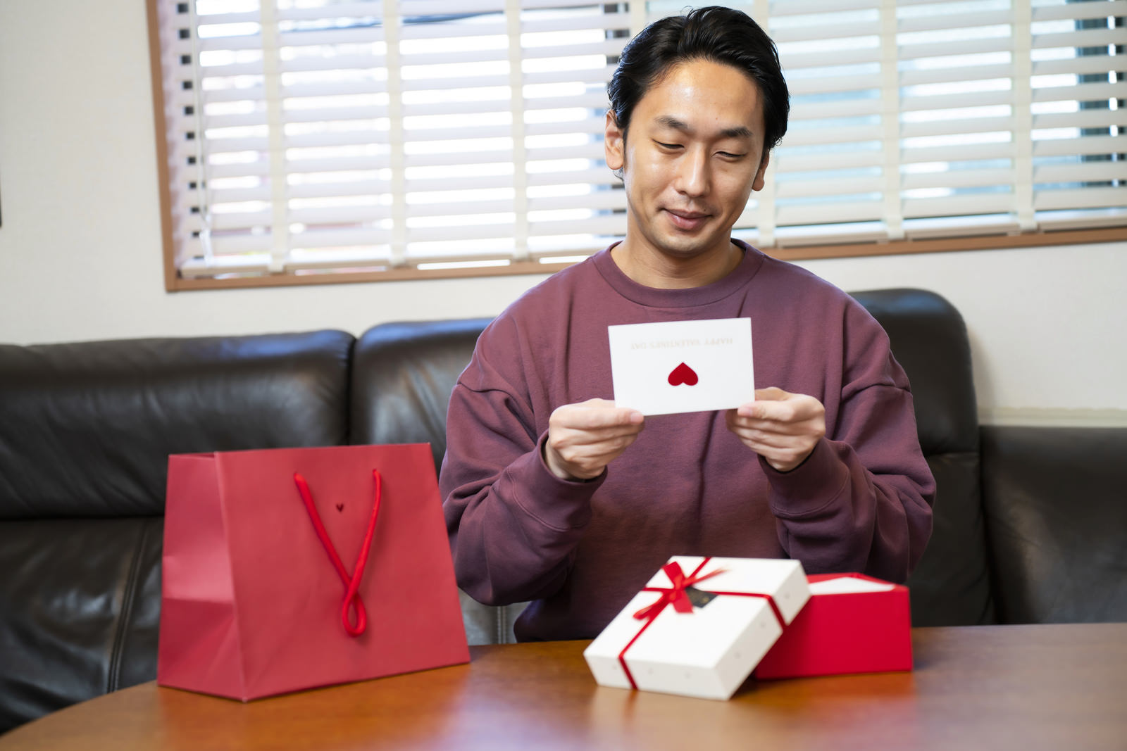 赤いギフトバッグとプレゼントボックスが置かれた机で、白い封筒のラブレターを持ってニヤニヤ笑う男性［モデル：大川竜弥］