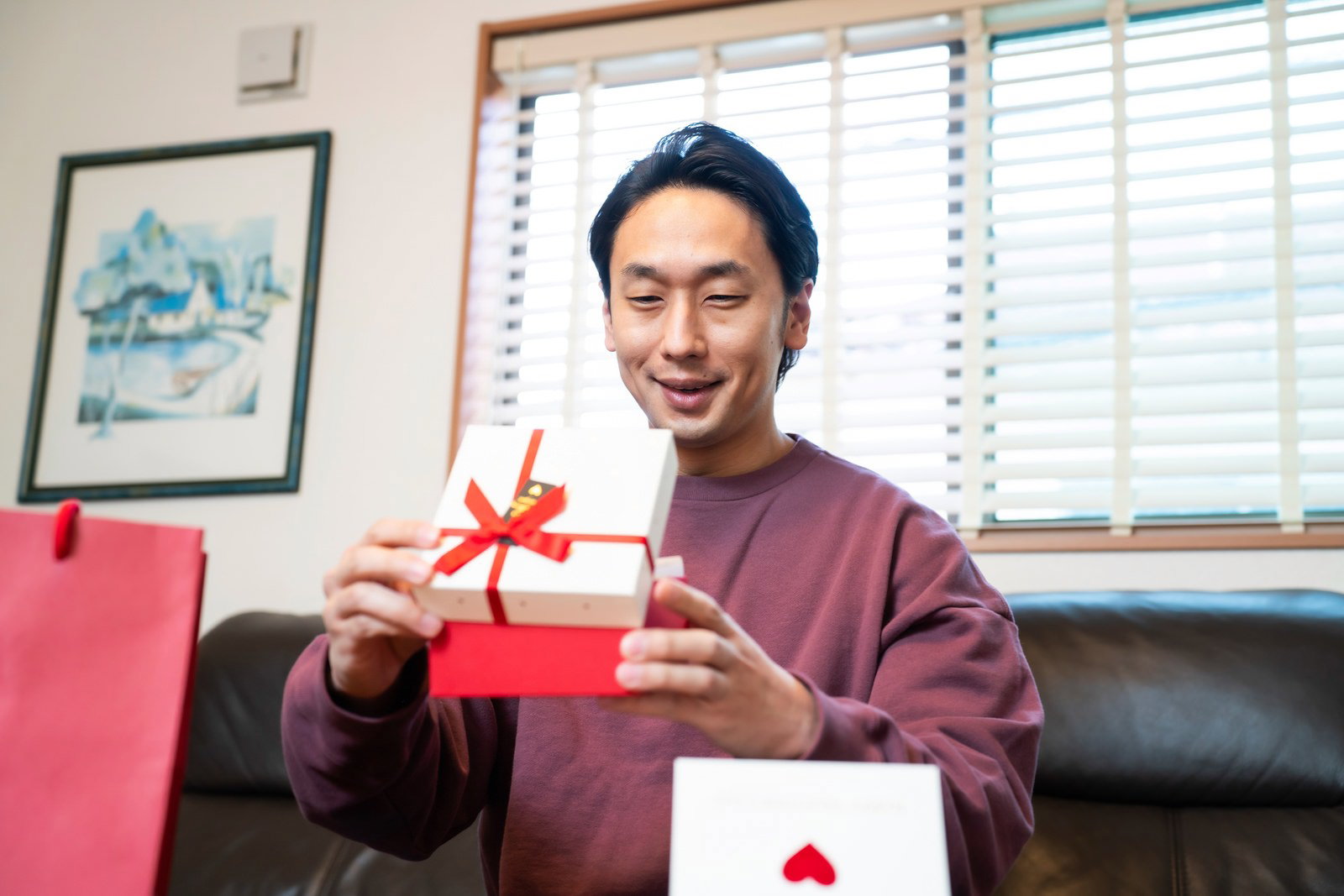 赤いリボンのプレゼントを両手で持ち、嬉しそうに笑う男性［モデル：大川竜弥］