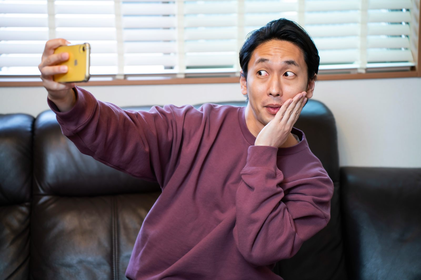 紫色のトレーナーを着た男性が顔に手を添えてスマートフォンで自撮りをしている［モデル：大川竜弥］