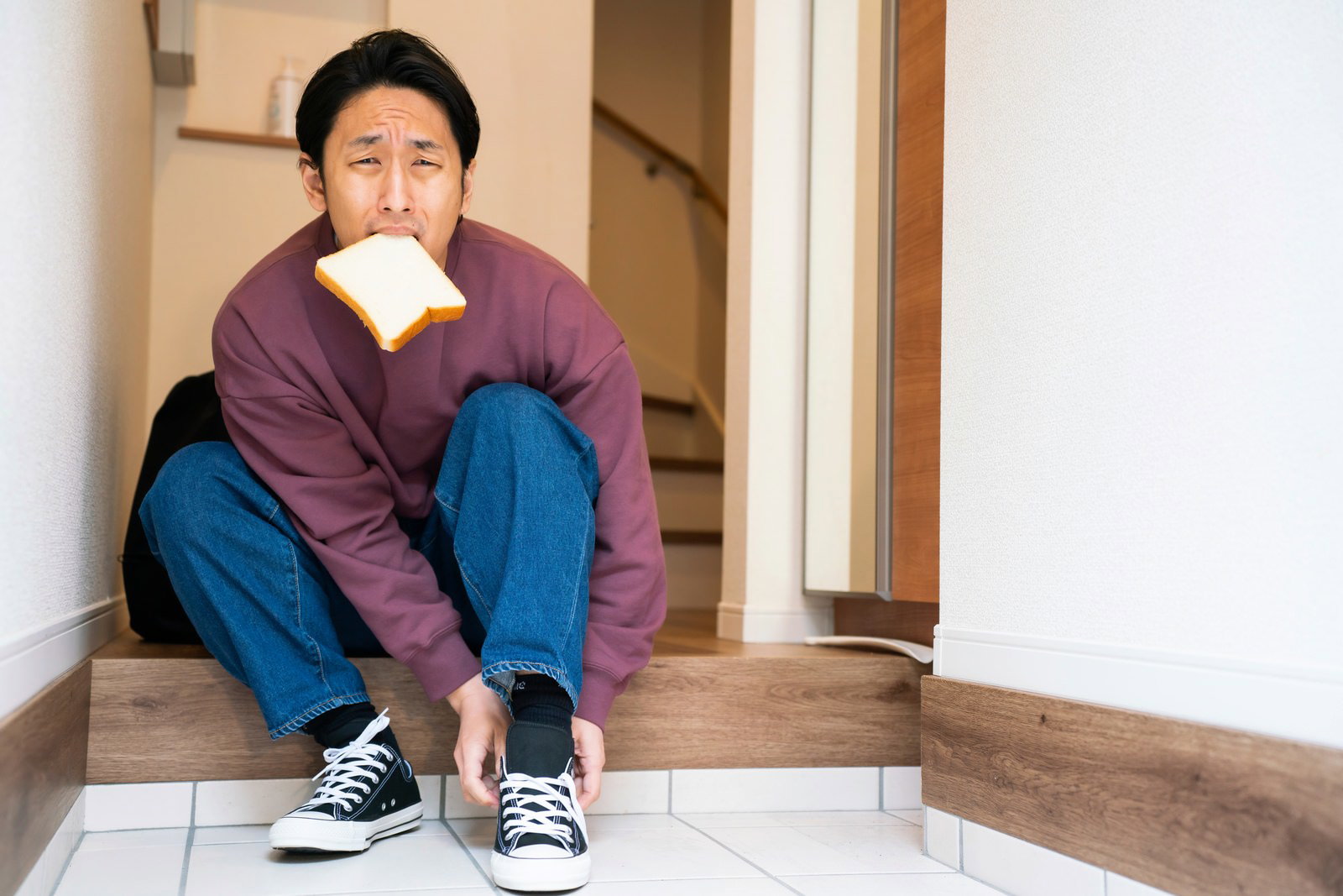 玄関で食パンを口にくわえながらスニーカーを履く男性［モデル：大川竜弥］