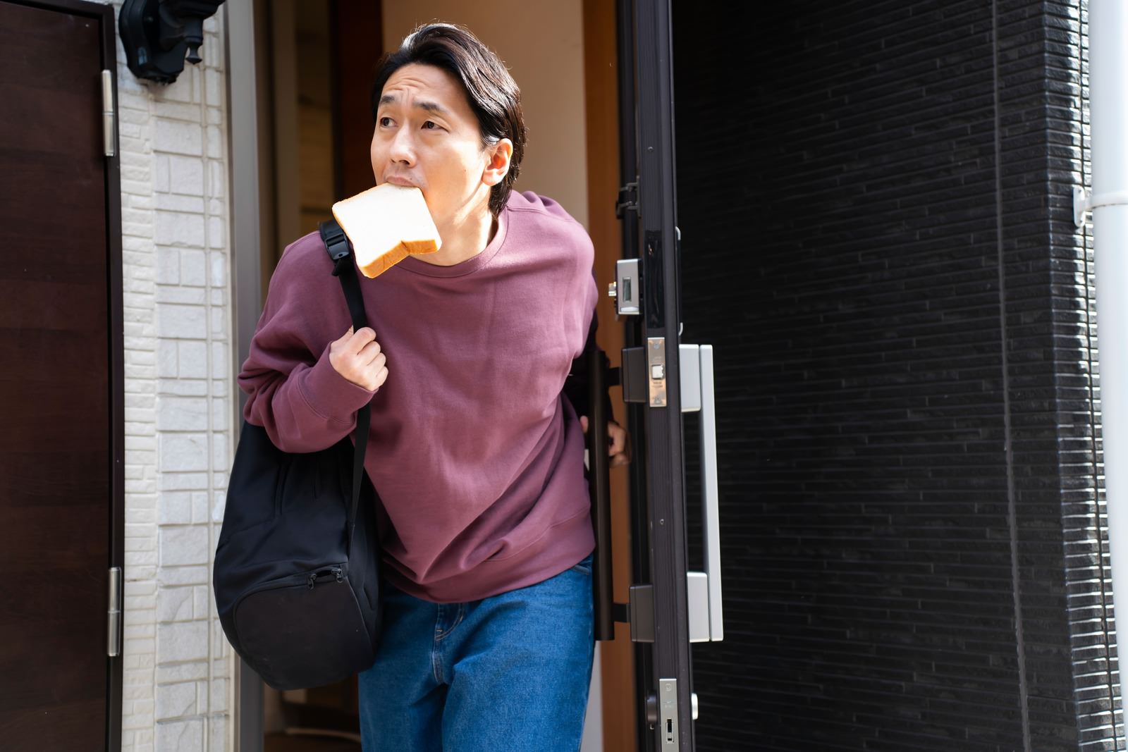 食パンを咥えて玄関から出る紫色のスウェットシャツの男性［モデル：大川竜弥］