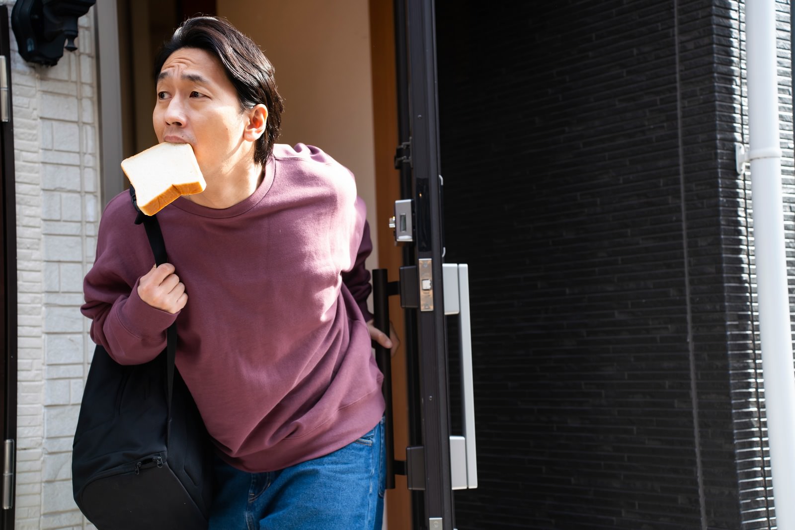 紫色の服を着た男性が玄関で食パンを咥えながら急いで外出している［モデル：大川竜弥］
