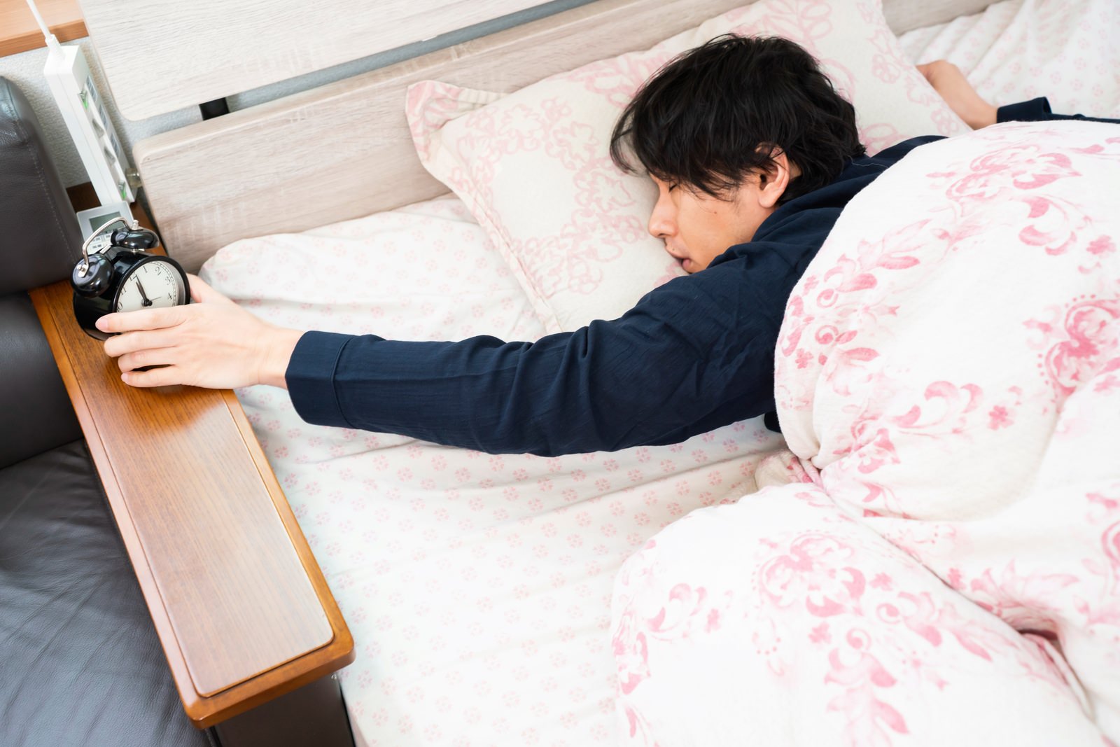 ベッドで横になりながら手を伸ばして目覚まし時計を止めようとする男性［モデル：大川竜弥］