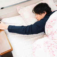 ベッドで寝ながら目覚まし時計を止める寝坊男子の写真