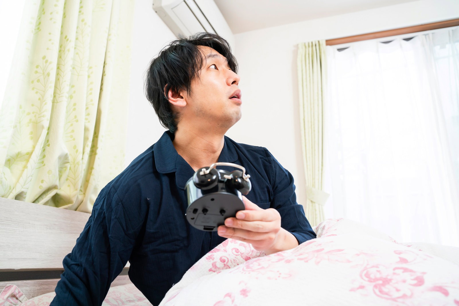 ベッドで目覚まし時計を持ち驚いた表情の男性［モデル：大川竜弥］