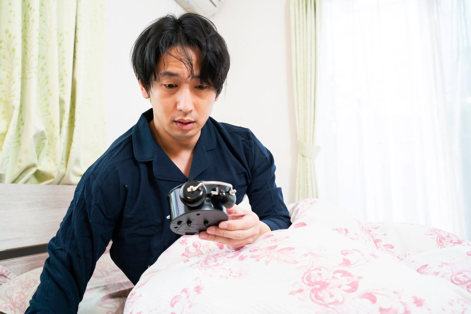 ベッドで横になりながら目覚まし時計を見つめる青いパジャマ姿の男性［モデル：大川竜弥］