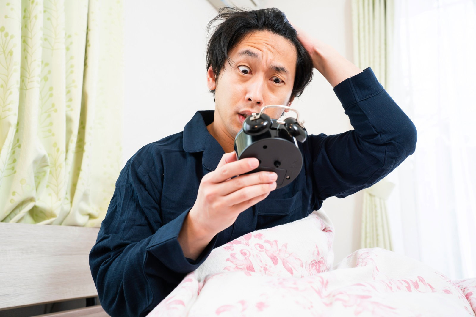 ベッドで頭に手を当ててスマートフォンを見ながら慌てている男性［モデル：大川竜弥］