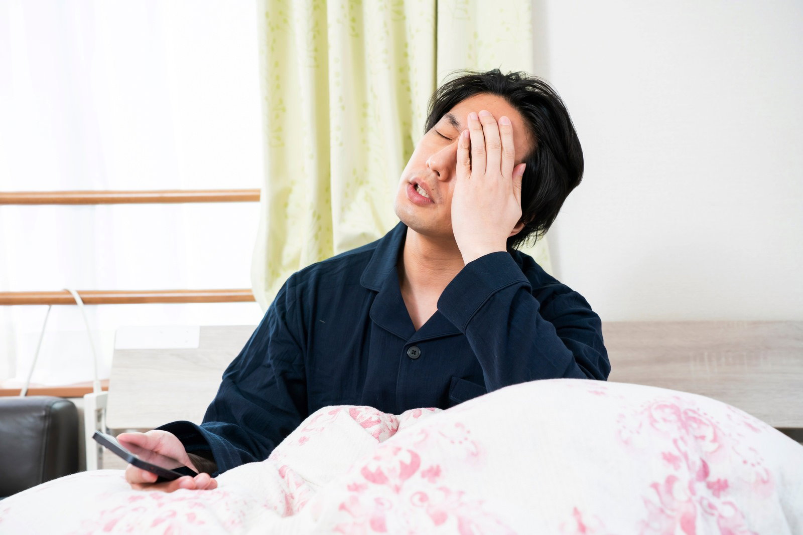 ベッドで頭を抱えながらスマートフォンを持つ男性［モデル：大川竜弥］