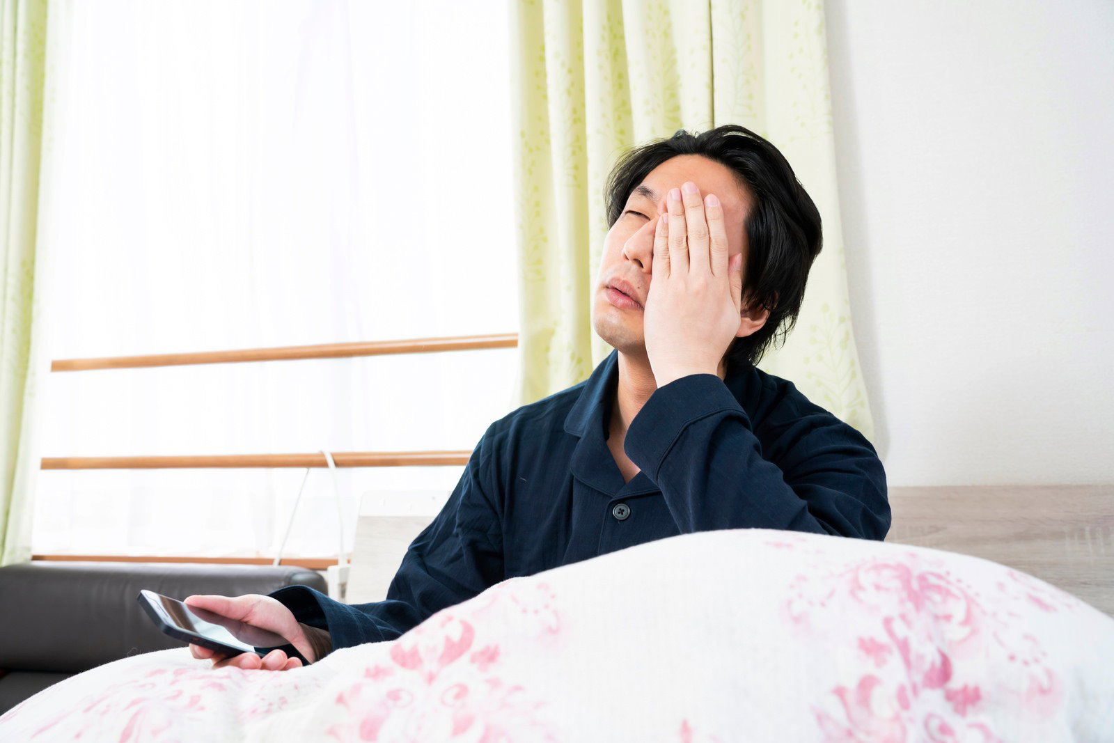 ベッドで寝坊して頭を抱えながらスマートフォンを見る男性［モデル：大川竜弥］