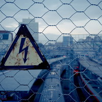 金網越しに見える錆びた高電圧危険標識と都市風景の写真