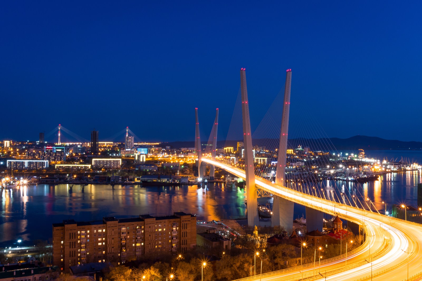 ロシア・ウラジオストクの夜景。2本の塔がある橋と光跡を描く車、港沿いの街灯に照らされた建物群