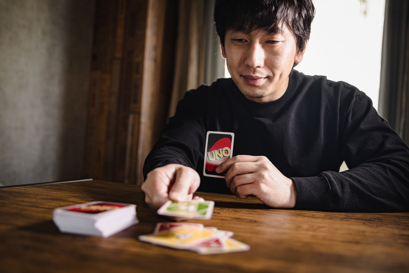 木製テーブルでUNOカードを手に持ち勝利目前の表情を見せる黒い長袖の男性［モデル：大川竜弥］