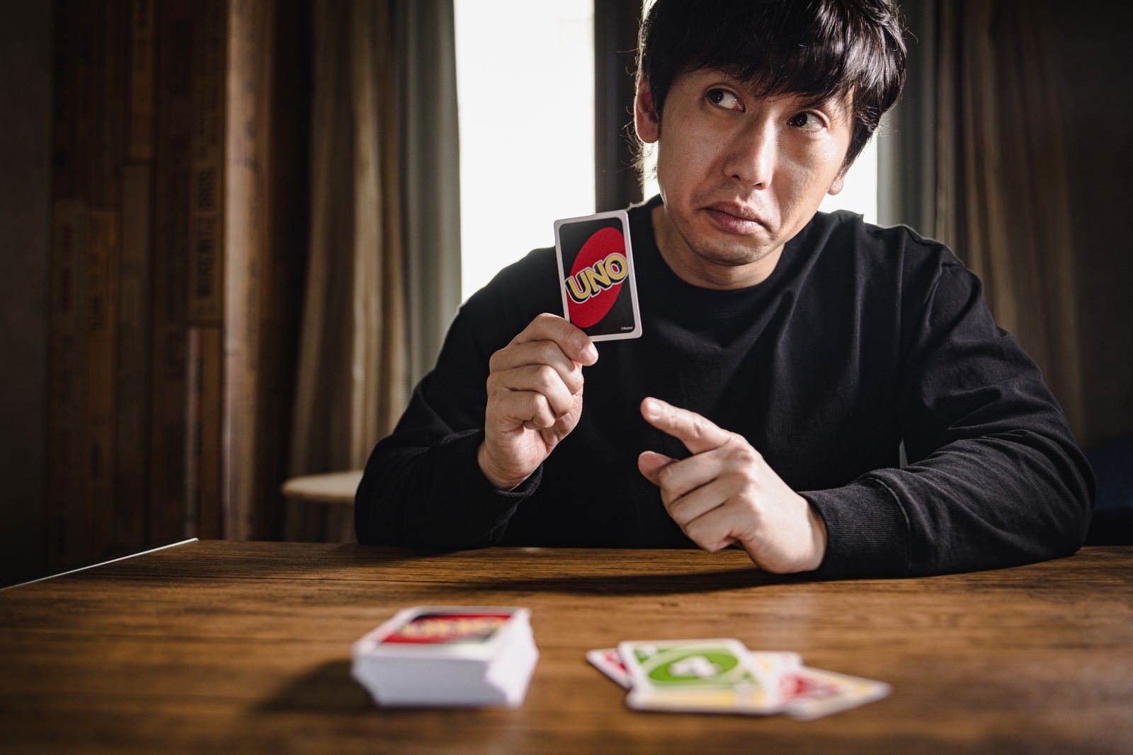 黒いセーター姿の男性がUNOカードを掲げ、相手を挑発する表情を見せている［モデル：大川竜弥］