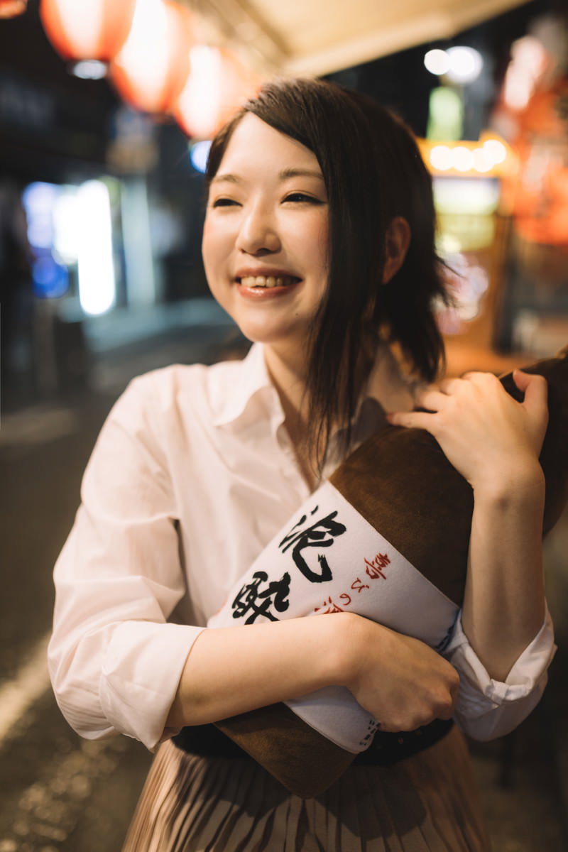 白いシャツを着た女性が泥酔と書かれた白地の抱き枕を抱えて、夜間の街で笑顔を向けている［モデル：シマヅ］