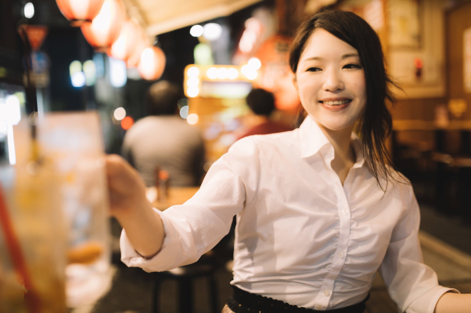 白いシャツを着た女性が右手を挙げて笑顔で乾杯している。背景は暖色系の照明が灯る夜間の飲食店［モデル：シマヅ］