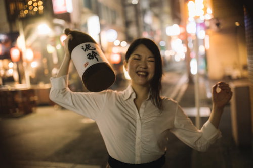 夜の繁華街で酒瓶を掲げて笑う酔っぱらいの女性