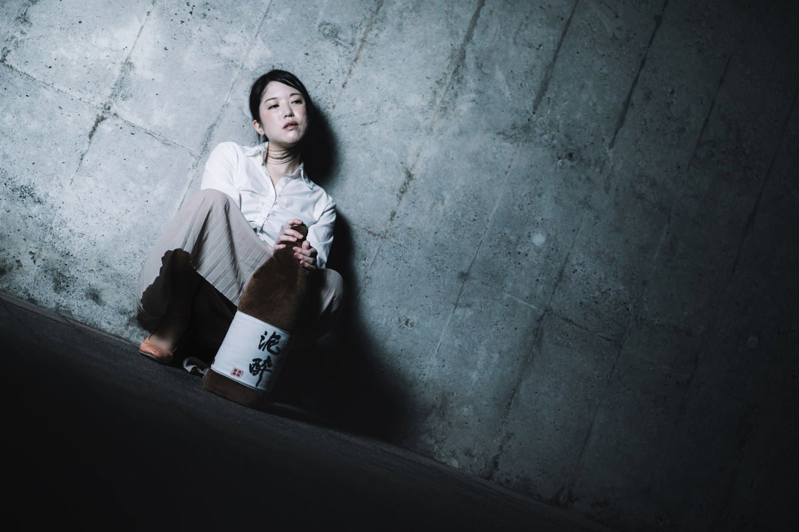 コンクリート壁に寄りかかってしゃがむ女性が日本酒の瓶を両手で抱えている［モデル：シマヅ］