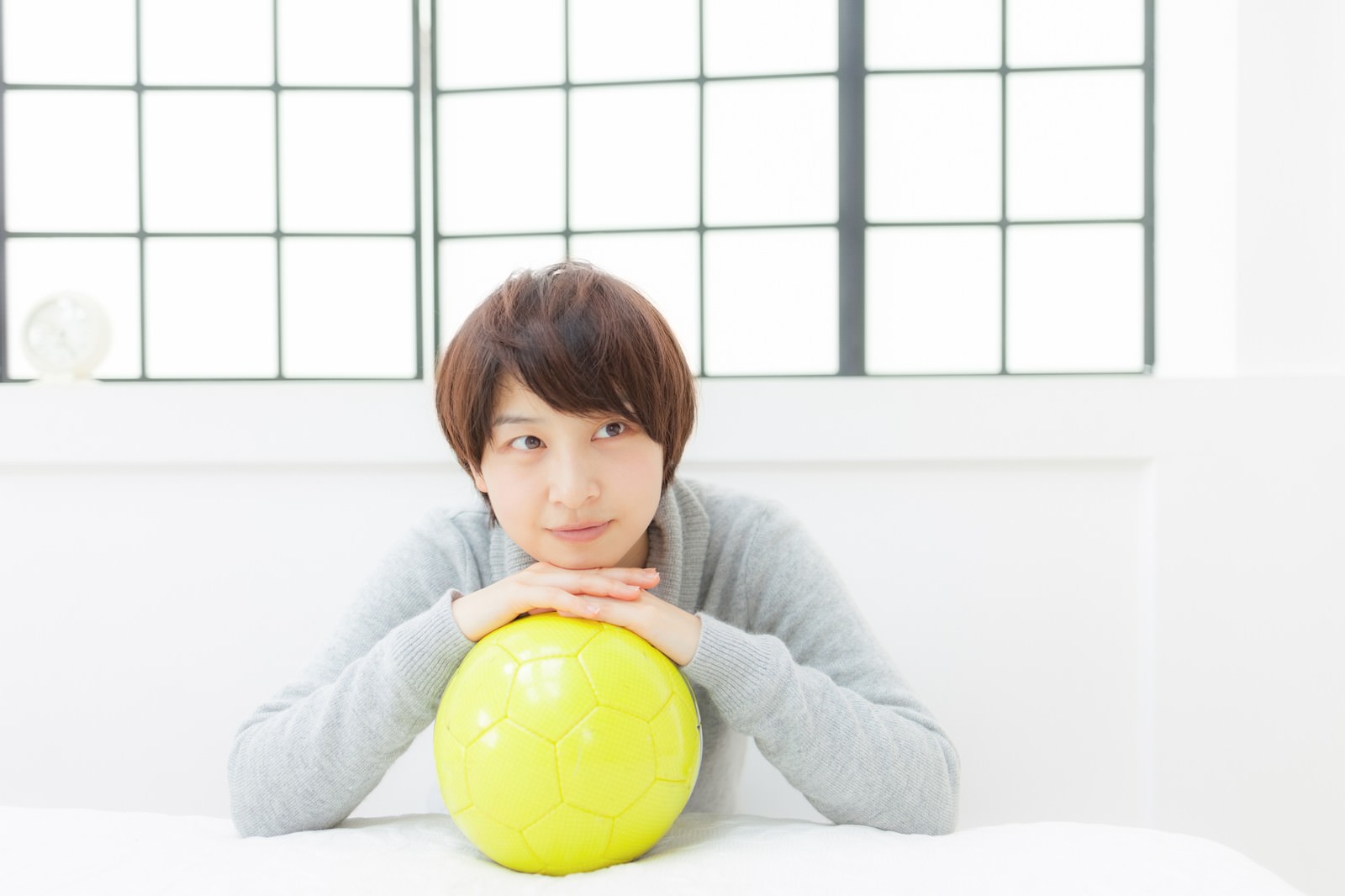 グレーのタートルネックを着た女性が黄色いサッカーボールを抱えている［モデル：八木彩香］
