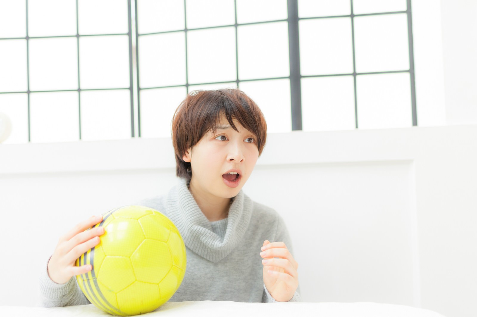 黄色いサッカーボールを持ちながら驚いた表情でテレビを見る女性［モデル：八木彩香］