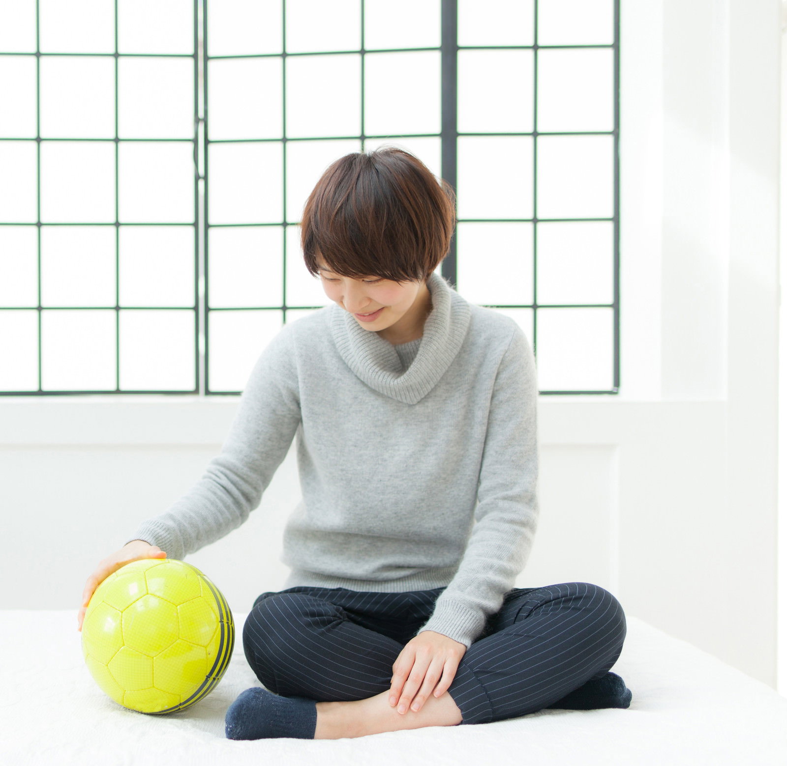 グレーのニットを着た女性が室内で床に座り、黄緑色のサッカーボールを持っている［モデル：八木彩香］