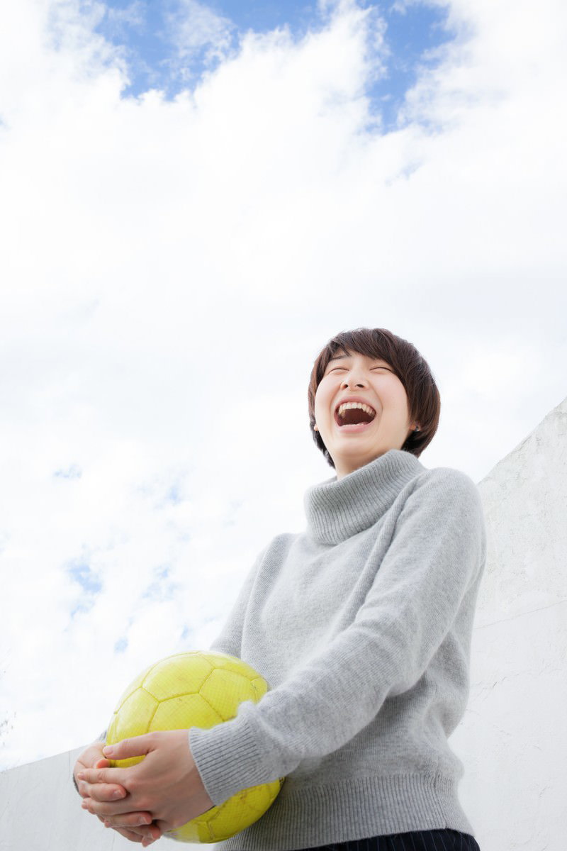 黄色いサッカーボールを持ち、青空を背景に大きく笑う女性［モデル：八木彩香］