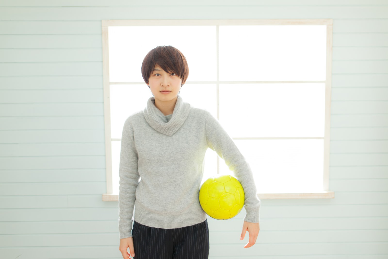 黄色いサッカーボールを持つ、グレーのタートルネックを着た女性コーチ［モデル：八木彩香］