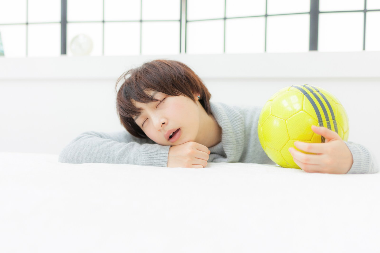 グレーのニットを着た女の子がソファーでサッカーボールを抱えて眠っている［モデル：八木彩香］