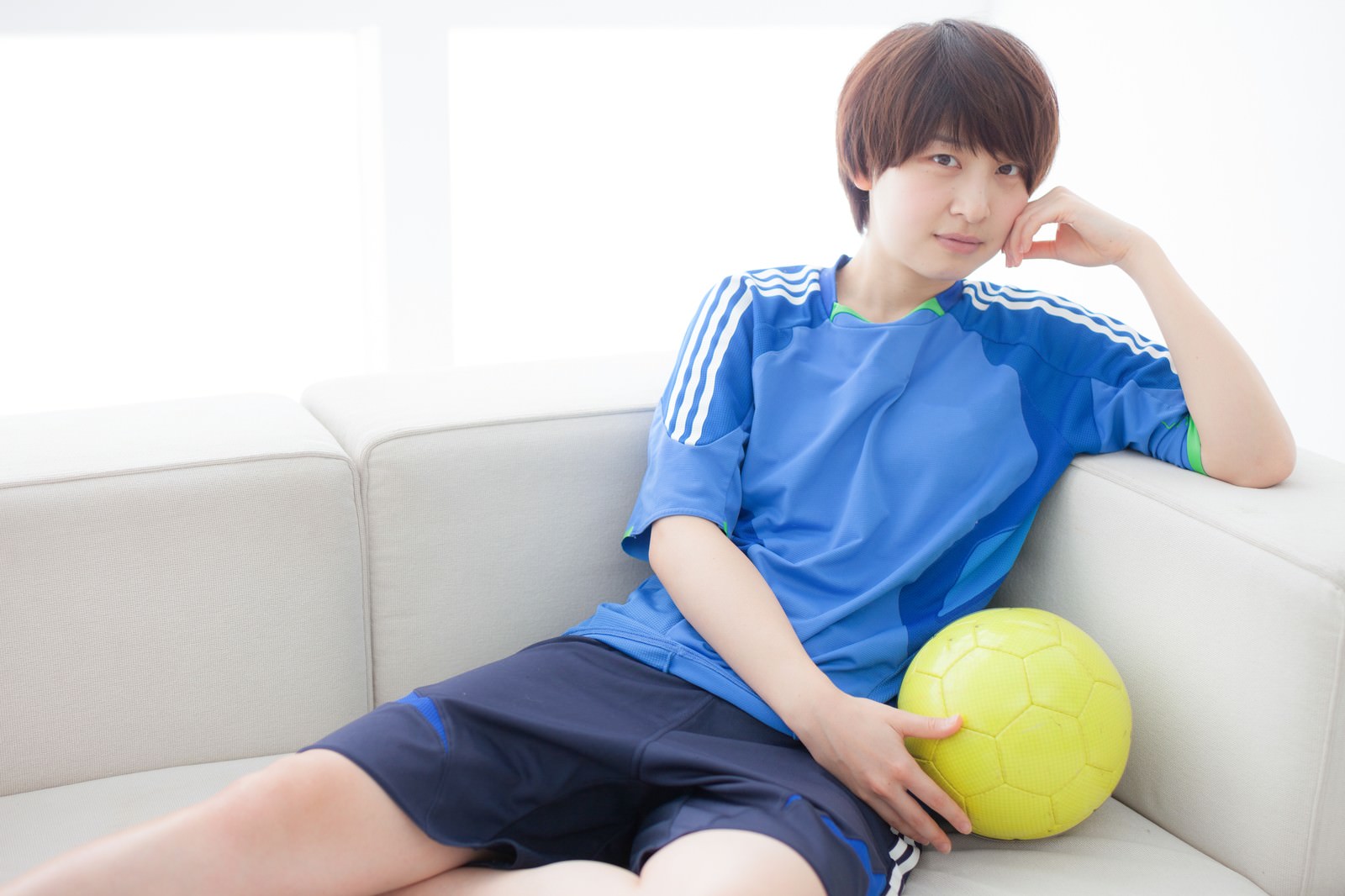 ソファーに座ってサッカーボールを抱える青いユニフォーム姿の女性［モデル：八木彩香］