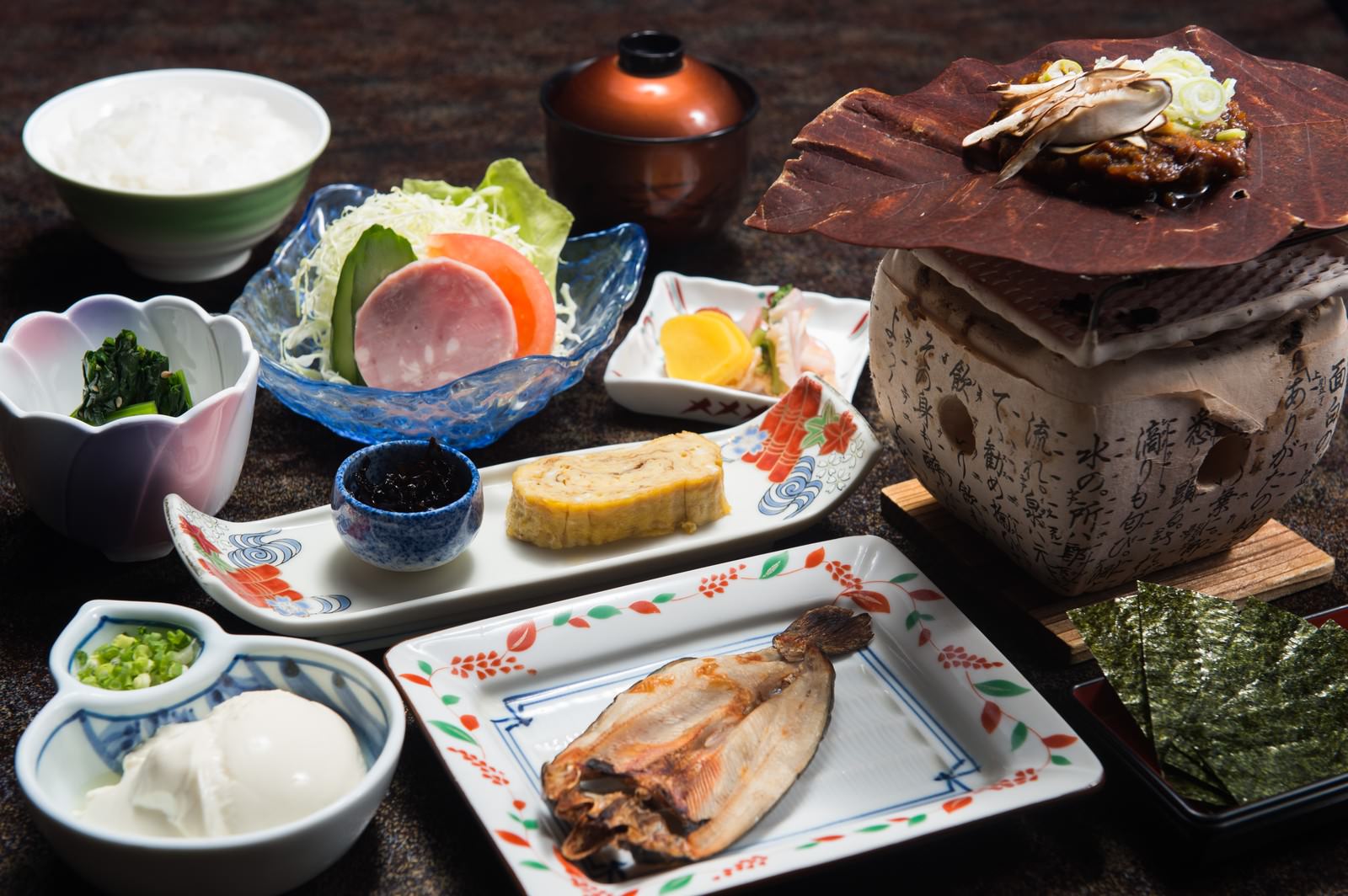 和朝食一式が黒い食卓に配置。白磁の小鉢や青絵食器、奥に土鍋、焼き魚と卵焼き