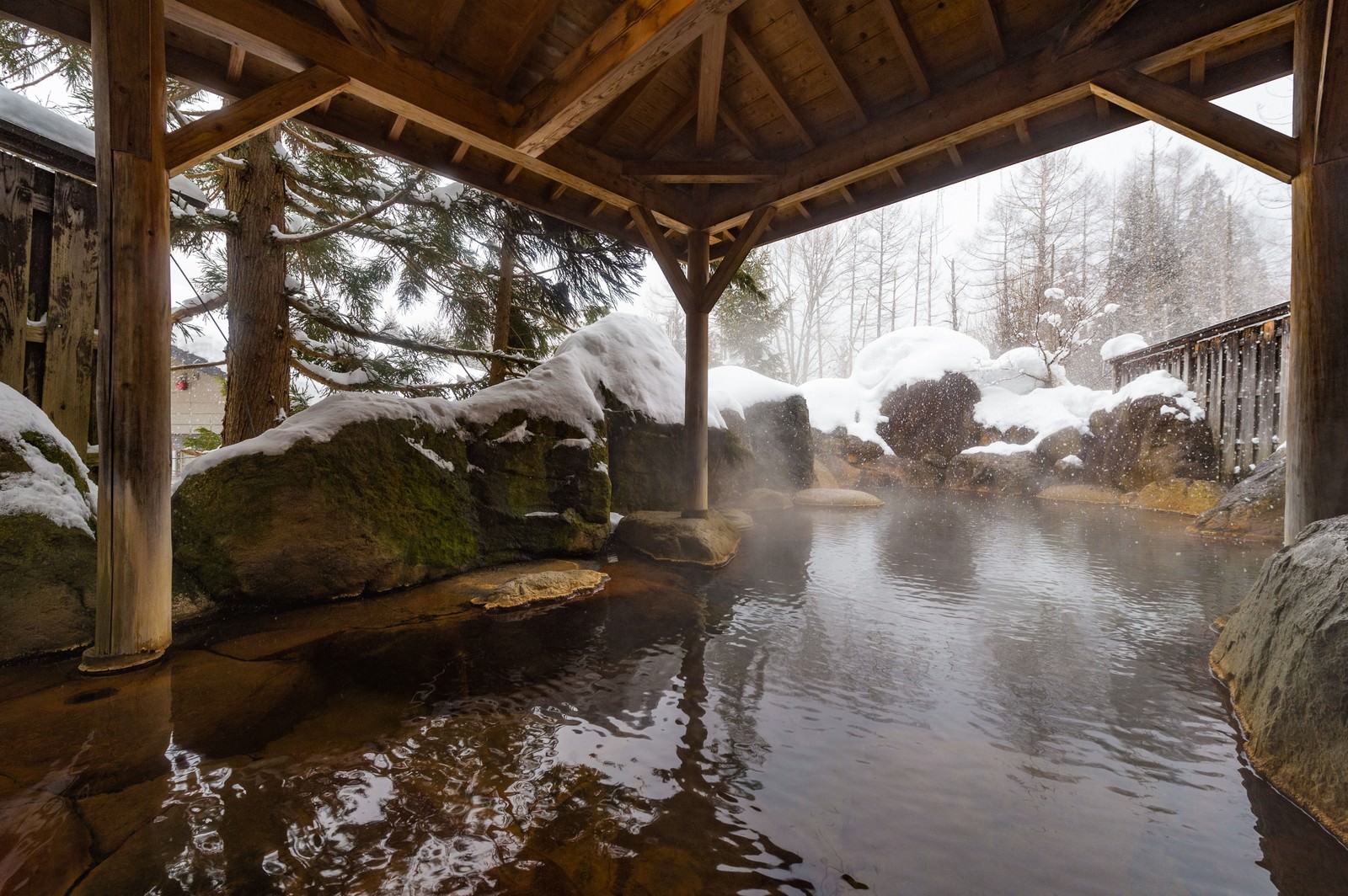 雪景色の中にある木造屋根付きの露天風呂から湯けむりが立ち上る［モデル：茜さや］