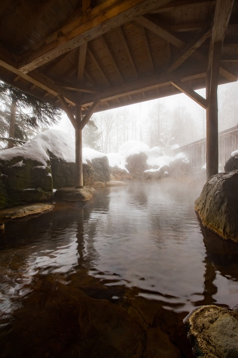 雪に覆われた木造屋根の露天風呂、湯けむりが立ち上る冬の温泉風景