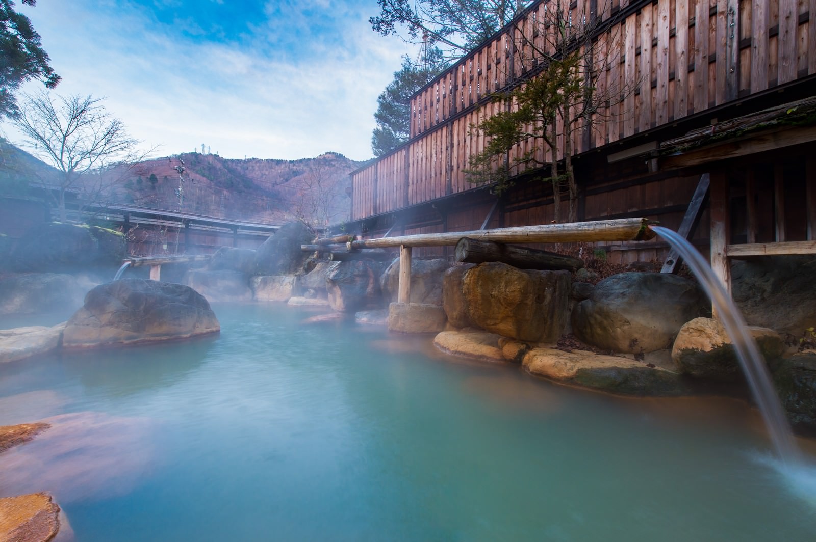 Stone-edged outdoor hot spring tub at Hirayu-kan with bamboo spout, steam rising, wooden building and autumn mountain ridge in background.