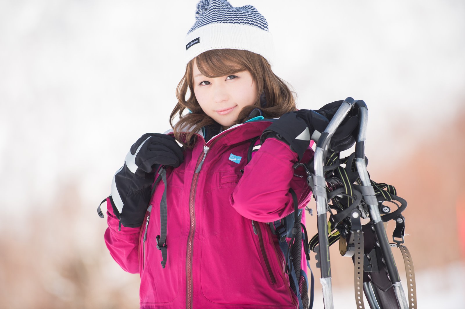 肩にスノーシューを担ぐピンク色ジャケット姿の女性が白背景で笑顔を向ける［モデル：茜さや］