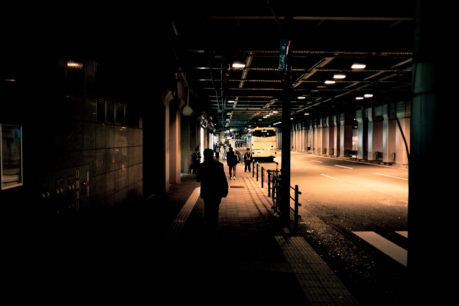 高架下の歩道でバス停留所に向かう人のシルエットが街灯に照らされている夜の風景