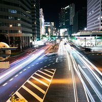 夜間の道路を直進する車の光跡と交差点の信号の写真