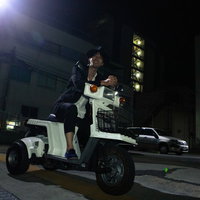 夜の街で原チャを乗り回す笑顔の男性、街灯に照らされた夜道での移動の写真