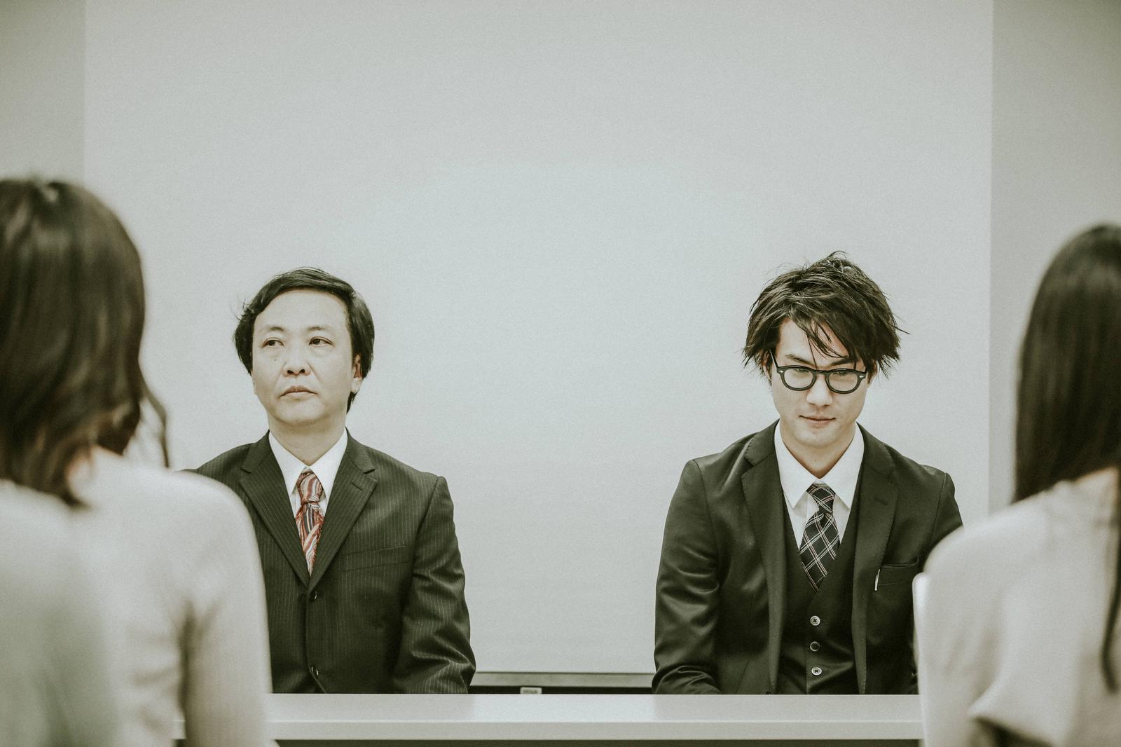 白いテーブルの前に座り記者の質問に応じる男性役員の記者会見の写真［モデル：のせさん 村田裕章］