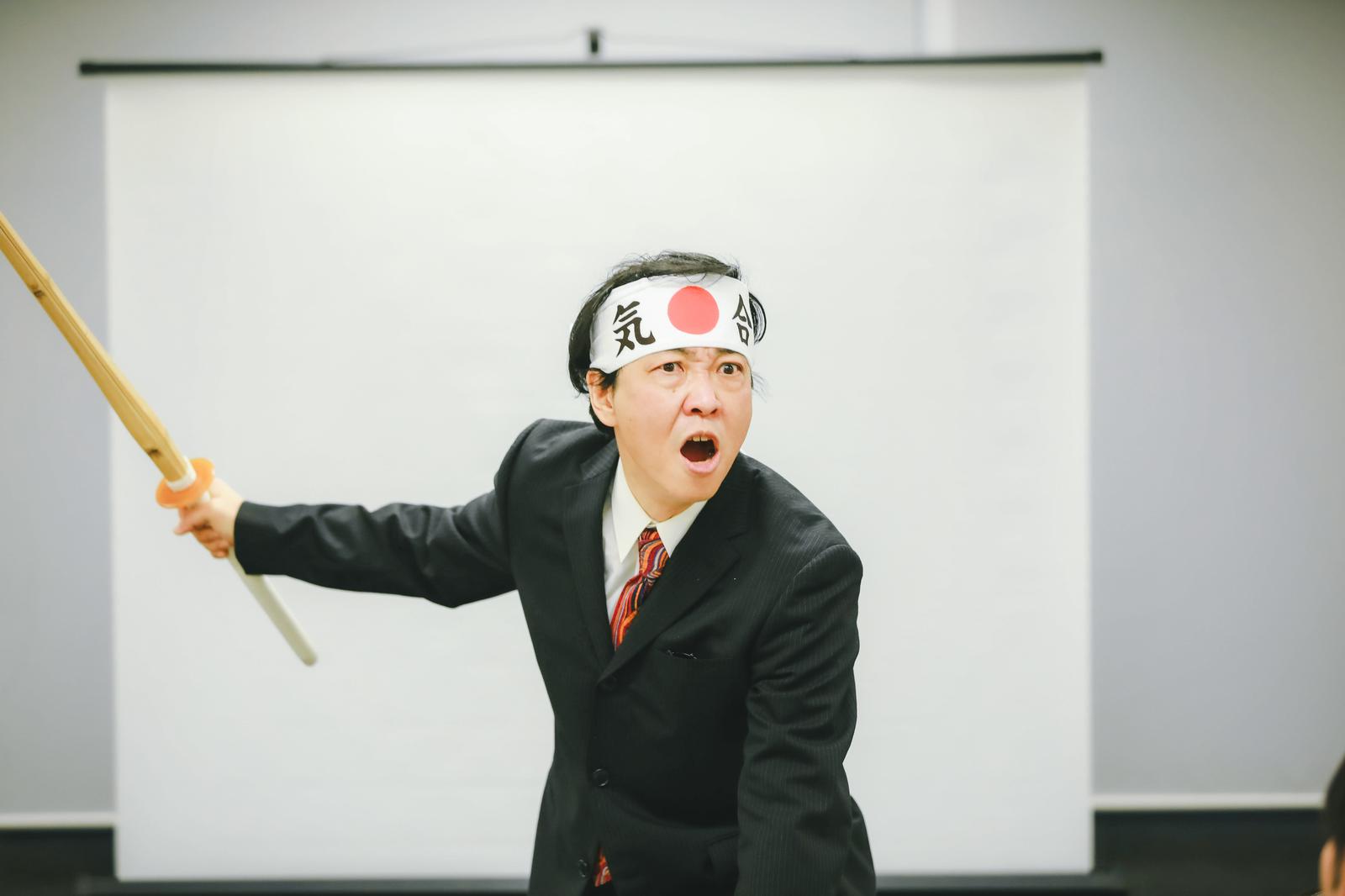 白いはちまきを巻いてスーツを着た男性が竹刀を握り気合を入れている［モデル：のせさん］